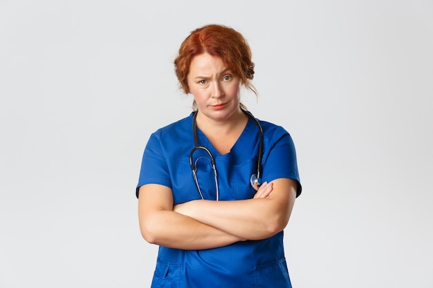 Medicine, healthcare and coronavirus concept. Skeptical and displeased medical worker, doctor looking with judgement at patient with raised eyebrow and crossed arms, standing grey background.