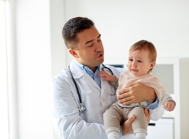 medicine, healthcare, pediatry and people concept - doctor or pediatrician holding sad crying baby girl on medical exam at clinic