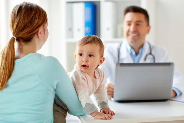 Photo medicine, healthcare, pediatry and people concept - happy woman with baby and doctor with laptop computer at clinic