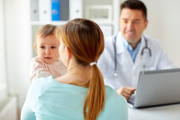 медицина, здравоохранение, педиатрия и концепция людей - счастливая женщина с ребенком и врач с ноутбуком в клинике