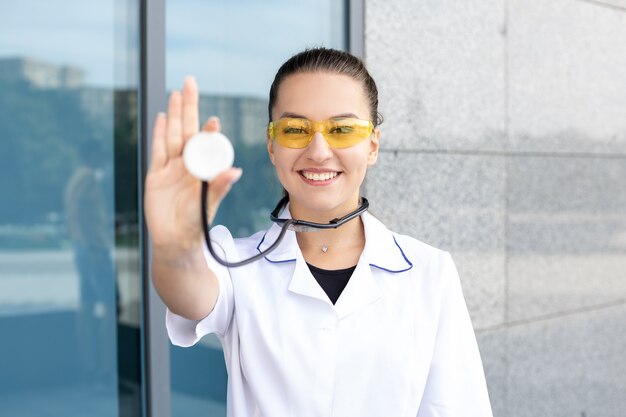 Medicine, healthcare, profession and people concept - European white female medic in white protective coat and yellow goggles shows stethoscope to camera outside in sunny weather