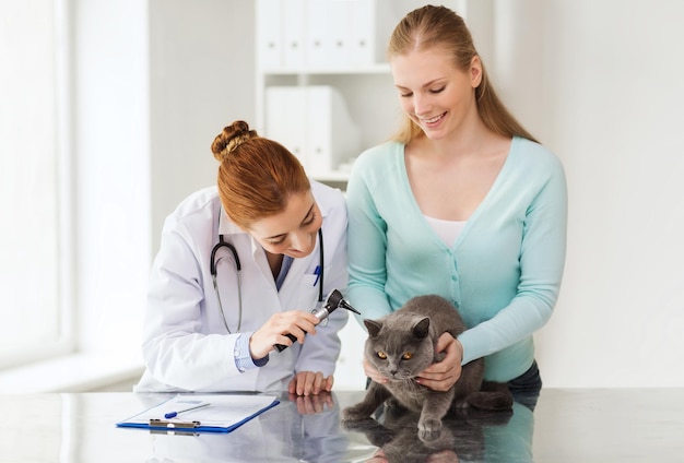 Concetto di medicina, animale domestico, animali, assistenza sanitaria e persone - donna felice e medico veterinario con otoscopio che controlla l'orecchio di gatto britannico presso la clinica veterinaria