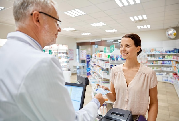 medicine, pharmaceutics, health care and people concept - smiling woman with wallet giving money to senior man pharmacist at drugstore cash register