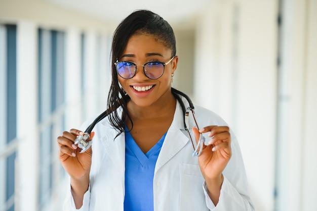 Концепция медицины, профессии и здравоохранения - счастливая улыбающаяся афро-американская женщина-врач в белом халате со стетоскопом на фоне больницы