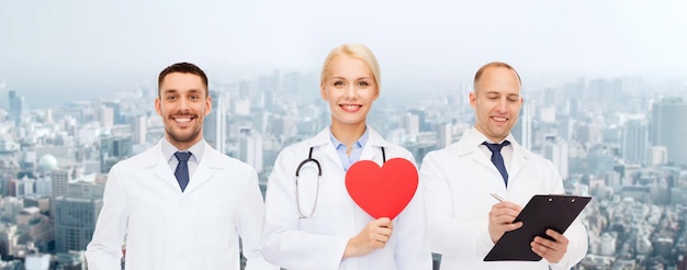 medicine, profession, teamwork and healthcare concept - group of smiling medics or doctors holding red paper heart shape, clipboard and stethoscopes over city background