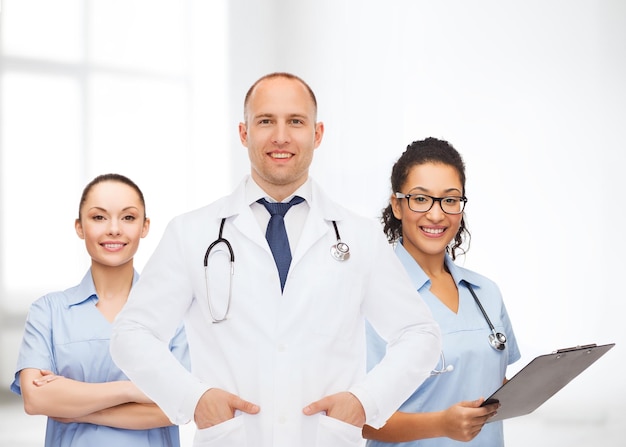 medicine, profession, teamwork and healthcare concept - international group of smiling medics or doctors with clipboard and stethoscopes over hospital background