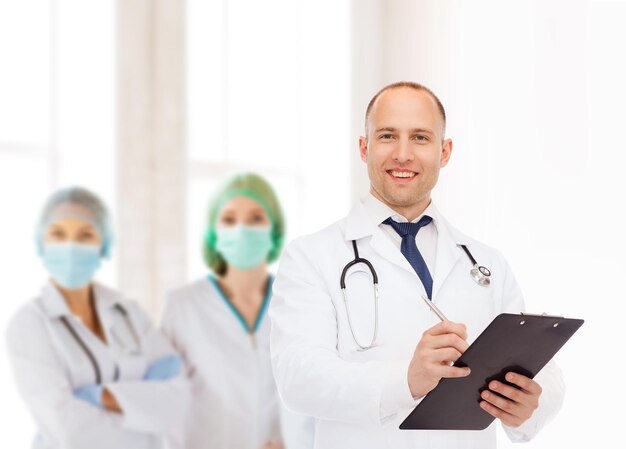 medicine, profession, teamwork and healthcare concept - smiling male doctor with clipboard and stethoscope writing prescription over group of medics