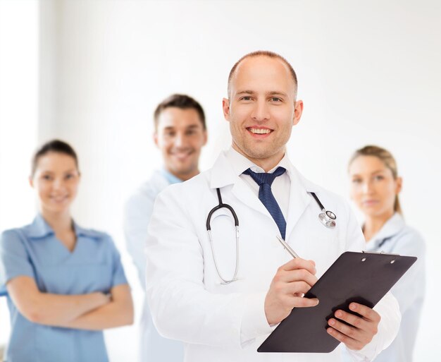 medicine, profession, teamwork and healthcare concept - smiling male doctor with clipboard and stethoscope writing prescription over group of medics