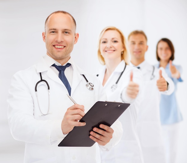医学、職業、チームワーク、ヘルスケアの概念-白い背景の上に処方箋を書くクリップボードと聴診器を持つ笑顔の男性医師