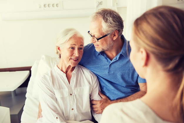 medicine, support, family health care and people concept - happy senior man and young woman visiting and cheering her grandmother lying in bed at hospital ward