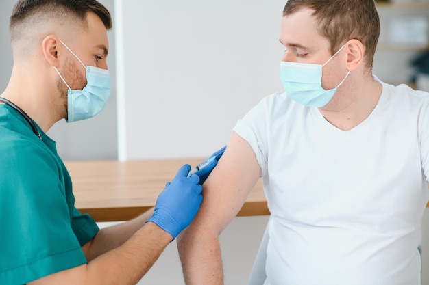 Vaccinazione medica e concetto di assistenza sanitaria medico che indossa una maschera medica protettiva per il viso per la protezione dalle malattie virali con siringa che esegue l'iniezione del vaccino a un paziente di sesso maschile