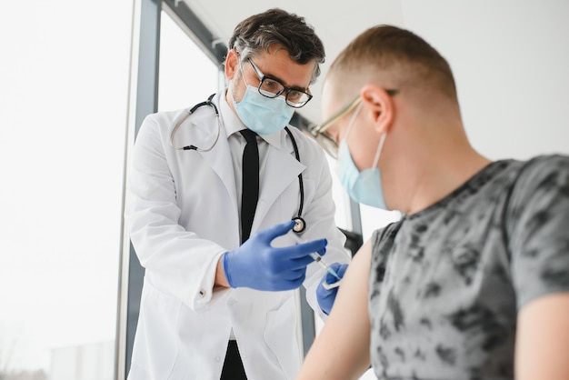 Photo medicine vaccination and healthcare concept doctor wearing face protective medical mask for protection from virus disease with syringe doing injection of vaccine to male patient