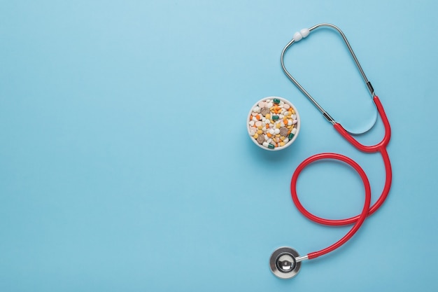 Medicines in a ceramic bowl and a red stethoscope on a blue background. Space for the text. Flat lay.