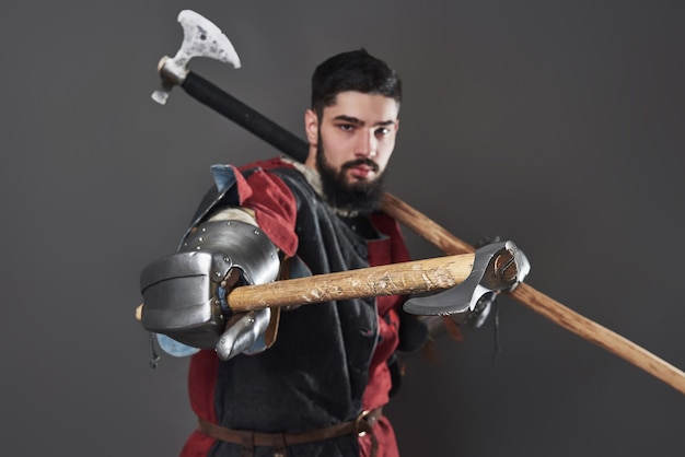 Medieval knight on grey background. Portrait of brutal dirty face warrior with chain mail armour red and black clothes and battle axe.