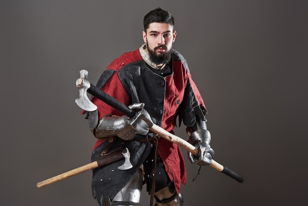 Medieval knight on grey. Portrait of brutal dirty face warrior with chain mail armour red and black clothes and battle axe