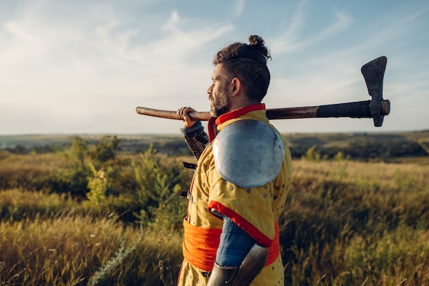 Medieval knight with axe poses in armor opposite the castle, great combat. Armored ancient warriors in armour posing in the field