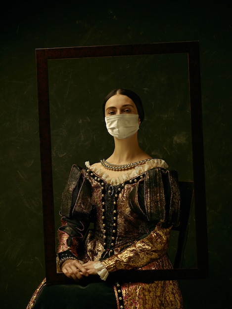 Средневековая молодая женщина в роли герцогини в защитной маске от распространения коронавируса на темно-синем фоне. Концепция сравнения эпох, здравоохранения, медицины и профилактики от пандемии.