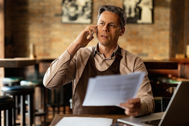 Medio volwassen ober die aan papierwerk werkt en op mobiele telefoon communiceert in een pub