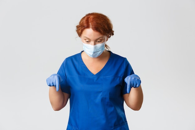 Medisch personeel, pandemie, coronavirus concept. portret van gelukkige arts bewondert iets naar beneden, vrouwelijke verpleegster in gezichtsmasker en handschoenen kijkt en wijst naar beneden met geïmponeerde uitdrukking