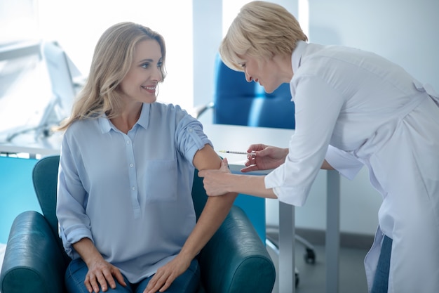 Medische injectie. Blonde vrouwelijke arts die injectie in de arm van glimlachende vrouwelijke patiënt maakt