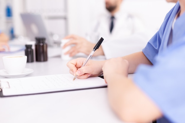 Medische vrouw beoefenaar schrijft notities op klembord blauw uniform dragen tijdens conferentie met collega's. Kliniek deskundige therapeut in gesprek met collega's over ziekte, medische professional.