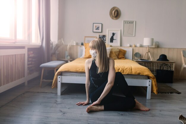 Meditating woman wearing a medical face mask to protect from corona virus, yoga position on mat home