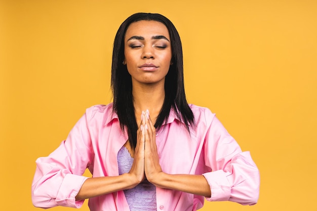 Photo meditation concept beautiful young african american woman stands in meditative pose enjoys peaceful atmosphere holds hands in praying gesture isolated over yellow background
