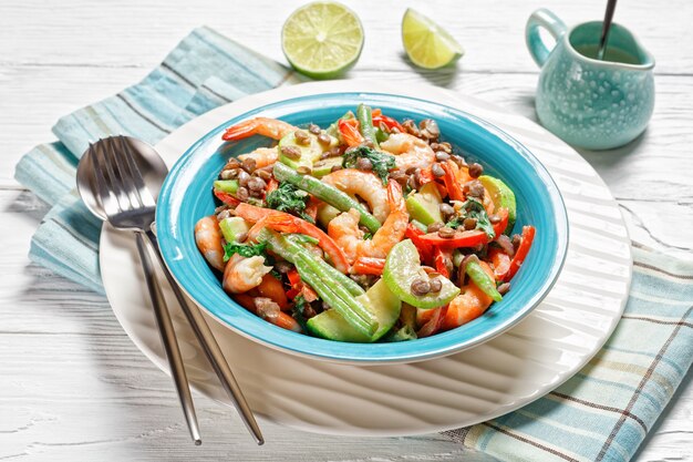 Mediterrane voorgerechtsalade van linzen, garnalen, sperziebonen, paprika's, met verwelkte boerenkool, limoen en tahinidressing geserveerd op een blauw bord op witte houten ondergrond, bovenaanzicht, close-up