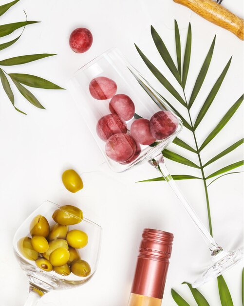 Foto composizione alimentare mediterranea con bicchieri di vino con uva e olive verdi bottiglia di vino rosa e foglie di palma su sfondo bianco il concetto di una località di vacanza estiva e rilassarsi