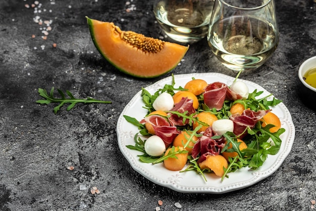 Mediterranean salad with prosciutto or jamon mozzarella and green basil leaves and Cantaloupe melon on dark table traditional Spanish and Italian appetizer served with wholemeal grissini top view