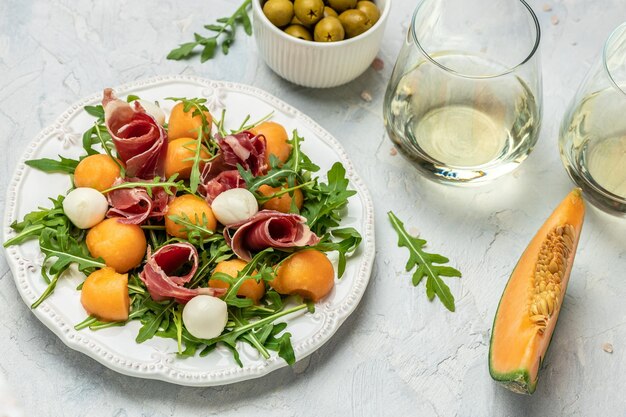 Mediterranean salad with prosciutto or jamon mozzarella and green basil leaves and Cantaloupe melon on white table traditional Spanish and Italian appetizer served with wholemeal grissini top view