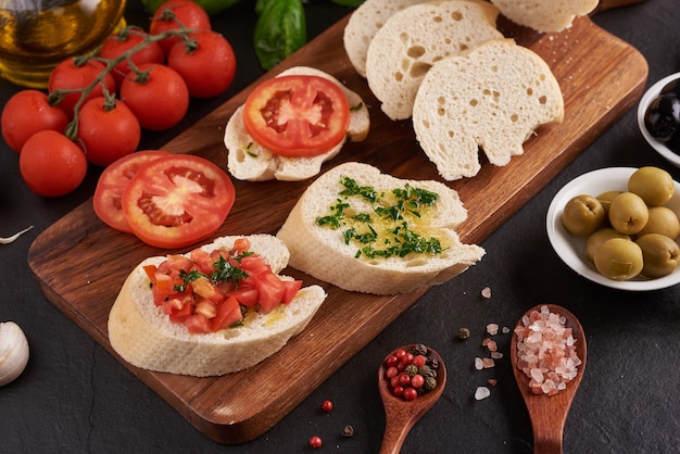 Mediterranean snacks set. Olives, oil, herbs and sliced ciabatta on a wooden board on black slate stone board over dark surface, juicy tomatoes on fresh bread, pesto as topping. top view. Flat lay