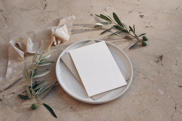 Mediterranean wedding stationery greeting card invitation mock\
up envelope on ceramic plate beige marble background olive tree\
fruit branches silk ribbons summer design top side view