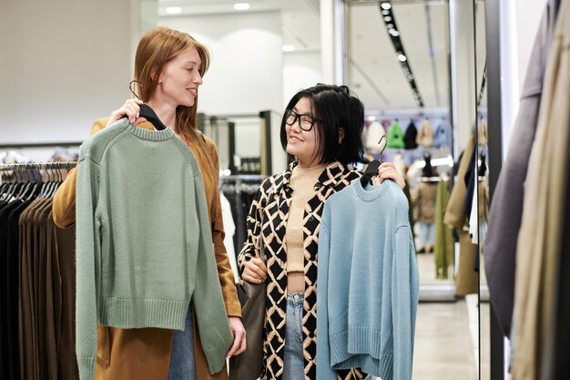 Photo medium long shot of two women showing clothes to each other while shopping at store