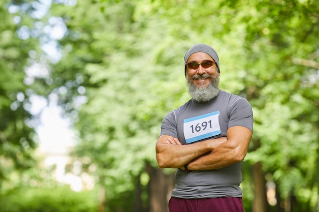 Medium portrait of joyful stylish senior male participant of summer marathon race standing with arms crossed smiling at camera, copy space