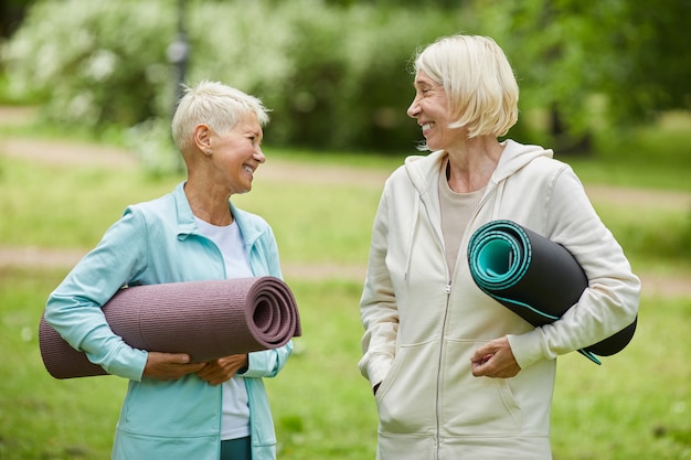 Medium portrait of two cheerful female friends wearing sports outfits holding yoga mats spending time together in park