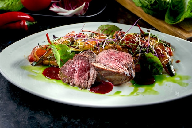 야채 장식을 곁들인 중간 희귀 쇠고기 메달리온이 흰색 접시에 제공됩니다. 어두운 대리석 테이블. 바베큐 고기, 레스토랑 음식. 맛있는 스테이크