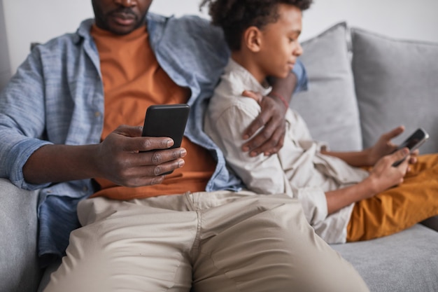 Medium section shot of unrecognizable African American man sitting on sofa with his teen son, both using Internet on smartphones