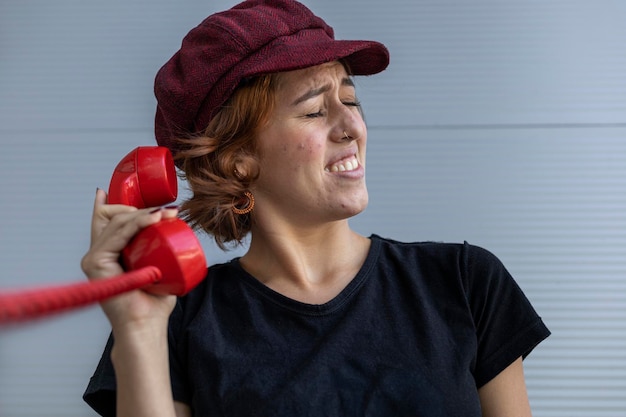 Medium short shot of latin american young woman 22 with cap and\
red hair uncomfortable because of the screams she hears from the\
conversation over the red retro handset vintage technology\
concept