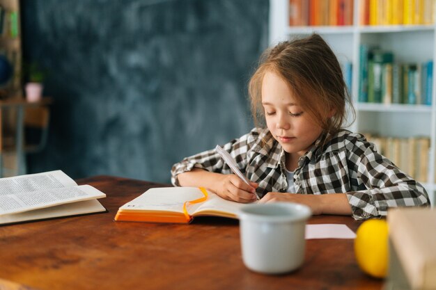 Medium shot of adorable pupil child school girl doing homework writing in notebook sitting at table