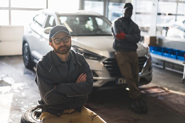 Medium shot of a caucasian auto mechanic sitting on wheels and an african mechanic leaning the car