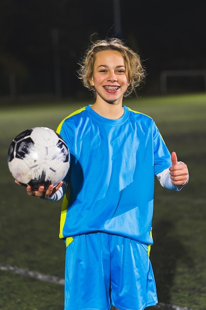 Medium shot of a cheerful little girl holding a soccer ball and showing a thumbup to a camera