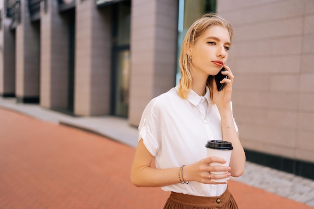 Medium shot of elegant blonde female teenager in casual clothes holding takeaway coffee cup and talking on smartphone during walking on city street