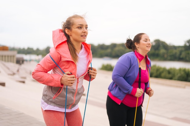 Medium shot of obese young woman doing squats exercises using fitness tape for weight loss with personal trainer outdoor in summer day