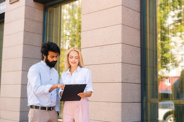 Medium shot portrait of attractive young blonde business woman explaining something on clipboard to handsome male business partner