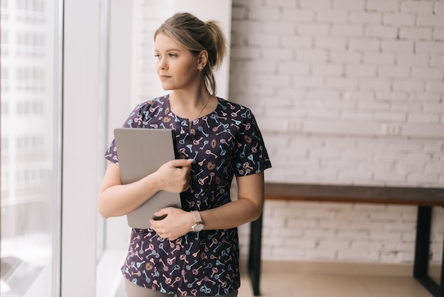 Medium shot portrait of attractive young woman office worker wearing casual clothing holding laptop computer in hands, standing by window and looking away