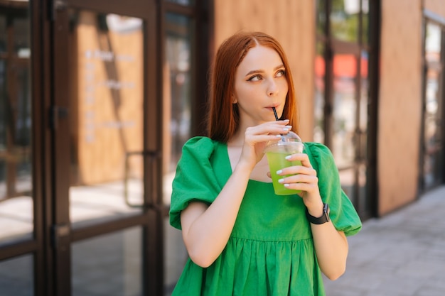 Medium shot portrait of cheerful attractive young woman holding cool cocktail standing at city street in sunny summer day, looking away. Pretty lady drinking cold beverage through straw.