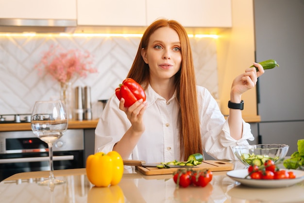 Medium shot portrait of cheerful redhead young woman holding in hand peppers sitting at wooden table