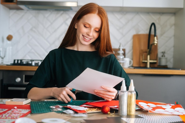 Medium shot portrait of happy young woman cutting out craft paper for envelopes with scissors making Christmas advent calendar on winter holidays eve