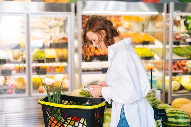 Medium shot portrait of pretty young woman checking products in grocery cart standing on background of shelves in supermarket